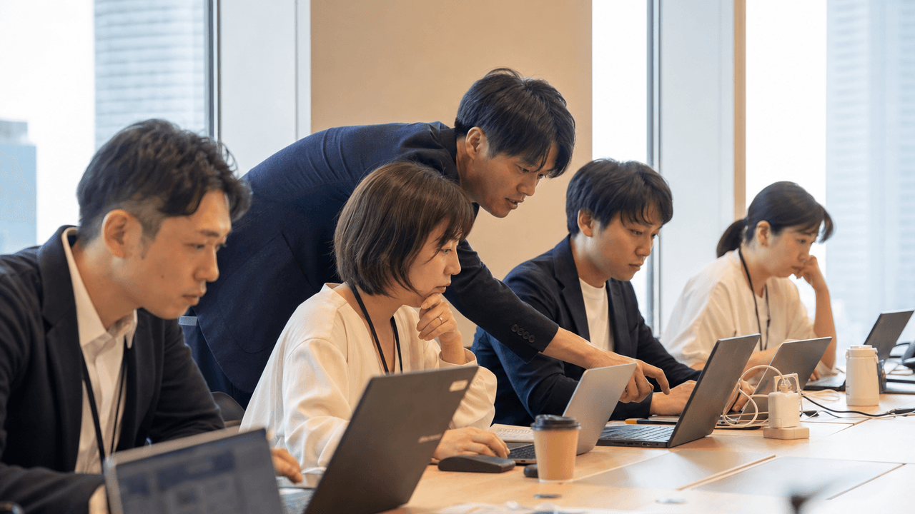 Day2 の会議室で各自がノートPCに集中し、講師が一人の画面を覗き込んで助言する様子
